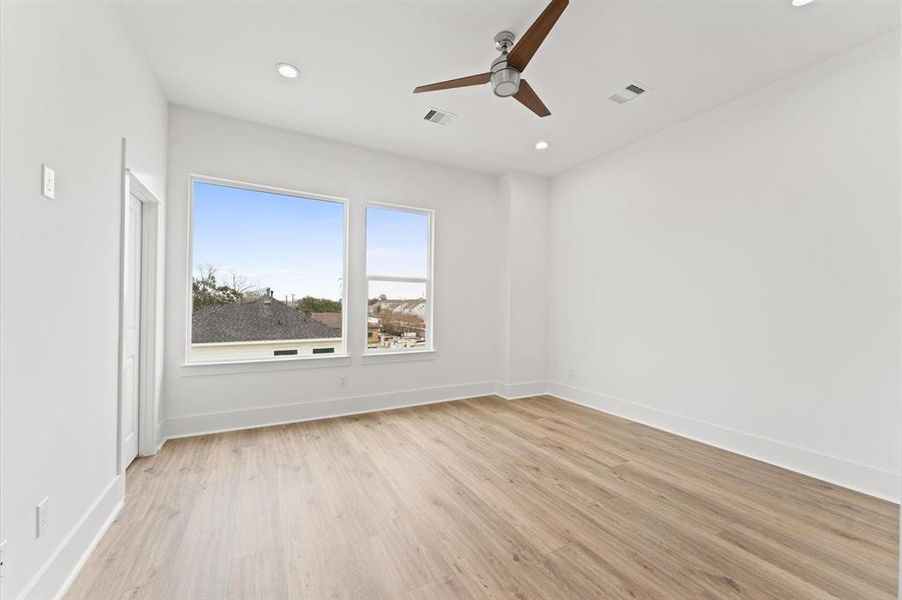 This spacious, bright bedroom features large windows with a pleasant view, light wood flooring, and a modern ceiling fan. The clean white walls and ceiling create a fresh and inviting atmosphere.