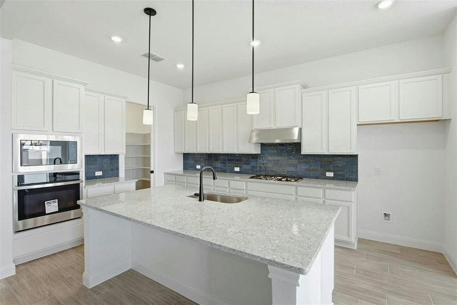 Spacious kitchen featuring a large central island with a sink and pendant lighting, white cabinetry, integrated stainless steel appliances, a dark tile backsplash, and wood-finish flooring