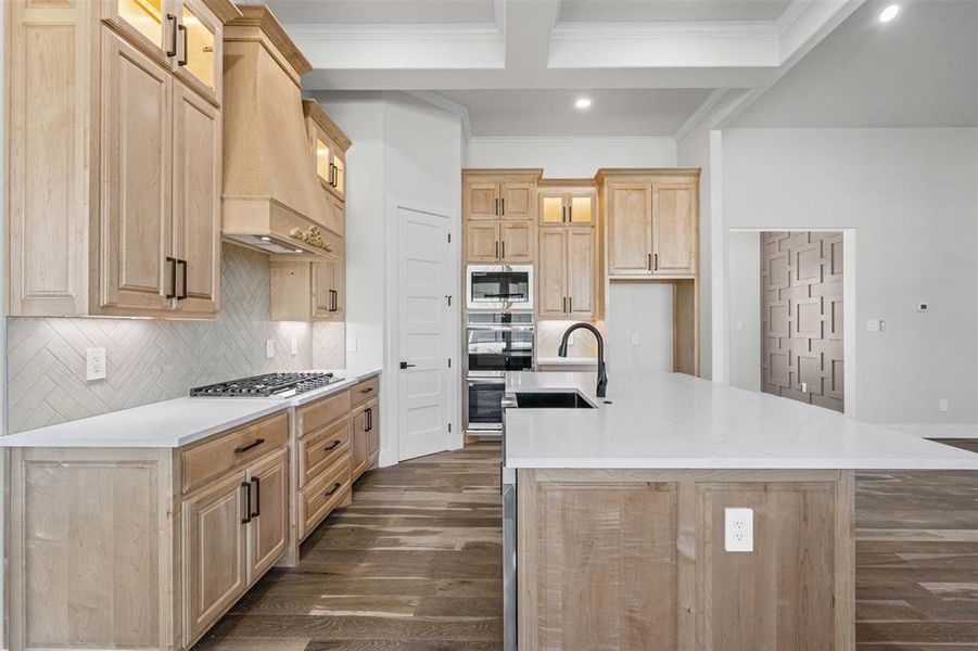 Kitchen featuring light brown cabinetry, light stone counters, tasteful backsplash, an island with sink, and beamed ceiling