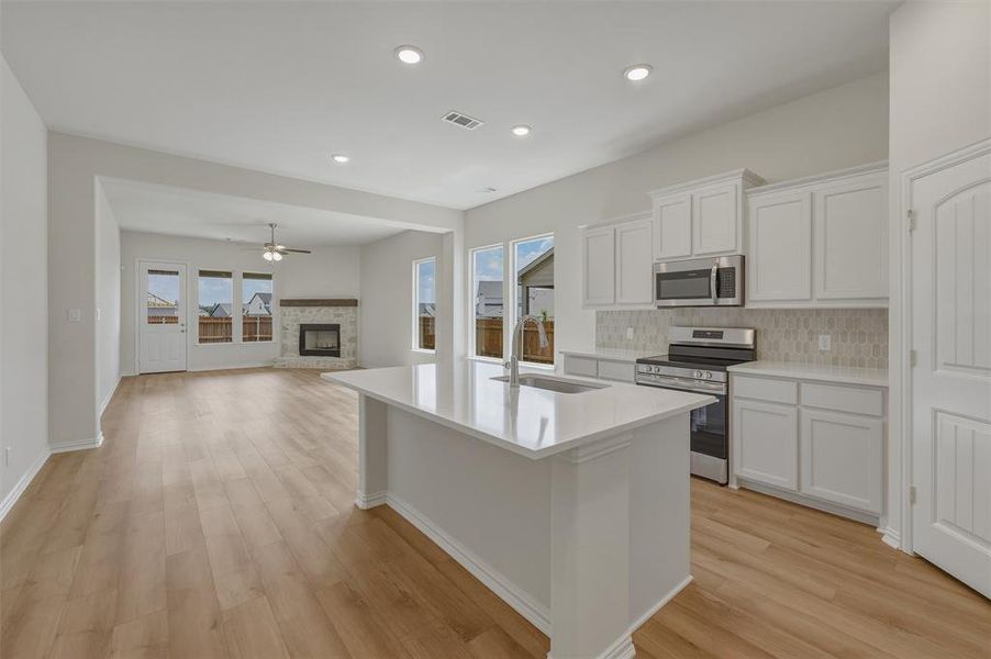 Open-concept living space featuring wood-finish flooring, a kitchen island with a sink, white cabinetry, stainless steel appliances, and a fireplace with a stone surround