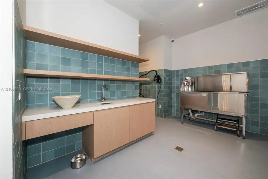 Utility space featuring custom wood cabinetry, quartz countertops, blue square tile wall backsplash, open floating shelving, and a stainless steel washing station