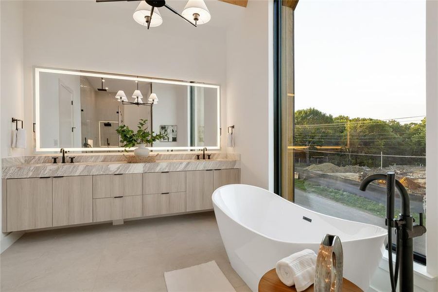 Modern bathroom featuring a freestanding soaking tub with floor-mounted fixture, a large vanity with dual sinks and light wood-finish cabinetry, a full-wall illuminated mirror, and a floor-to-ceiling window Modern bathroom featuring a freestanding soaking tub with floor-mounted fixture, a large vanity with dual sinks and light wood-finish cabinetry, a full-wall illuminated mirror, and a floor-to-ceiling window