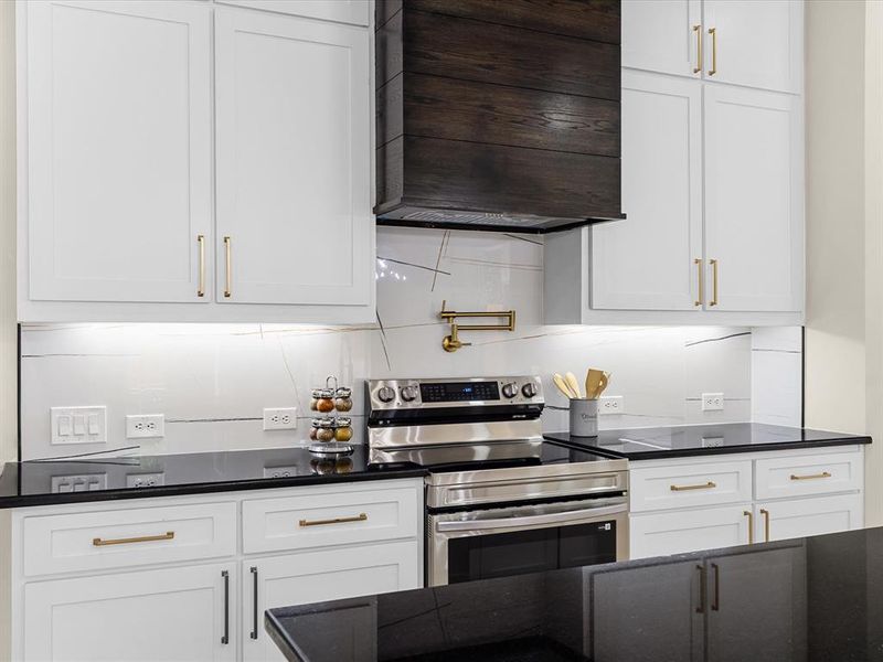 Modern kitchen featuring white shaker cabinetry with gold-finish hardware, a wood-finish range hood, stainless steel electric range, and dark countertops