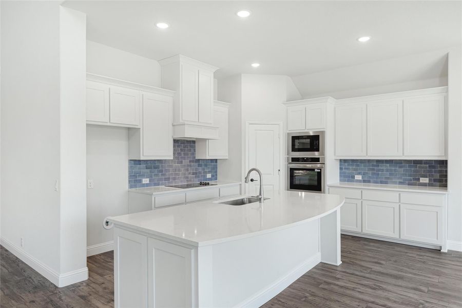 Kitchen with white cabinets, blue tile backsplash, curved island with sink, and stainless steel wall ovens