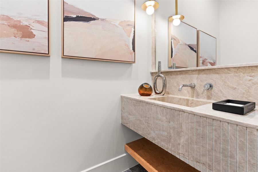Designer half bath featuring a floating stone vanity with integrated sink, wall-mounted chrome fixtures, textured detailing, warm brass sconces, and natural wood accent shelf. Designer half bath featuring a floating stone vanity with integrated sink, wall-mounted chrome fixtures, textured detailing, warm brass sconces, and natural wood accent shelf.