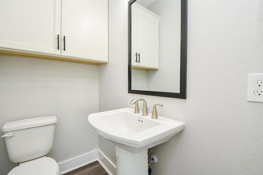 This is a compact and modern half-bathroom featuring a white pedestal sink with brushed nickel fixtures, a framed mirror above, and a white toilet. There's also an overhead cabinet for storage, and the room has neutral-colored walls and flooring, presenting a clean and bright space.