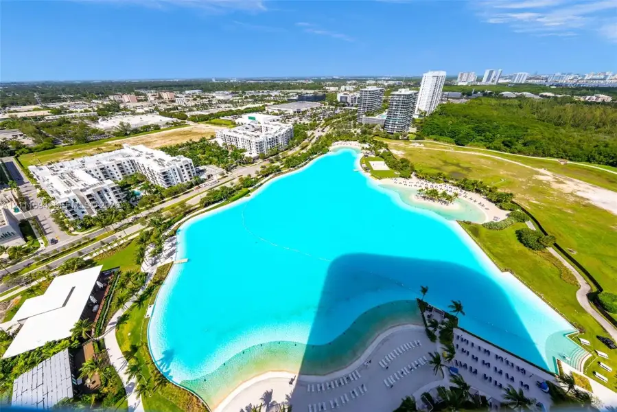 Aerial perspective featuring a large crystalline lagoon, expansive sandy shoreline, manicured green space, and paved pathways Aerial perspective featuring a large crystalline lagoon, expansive sandy shoreline, manicured green space, and paved pathways