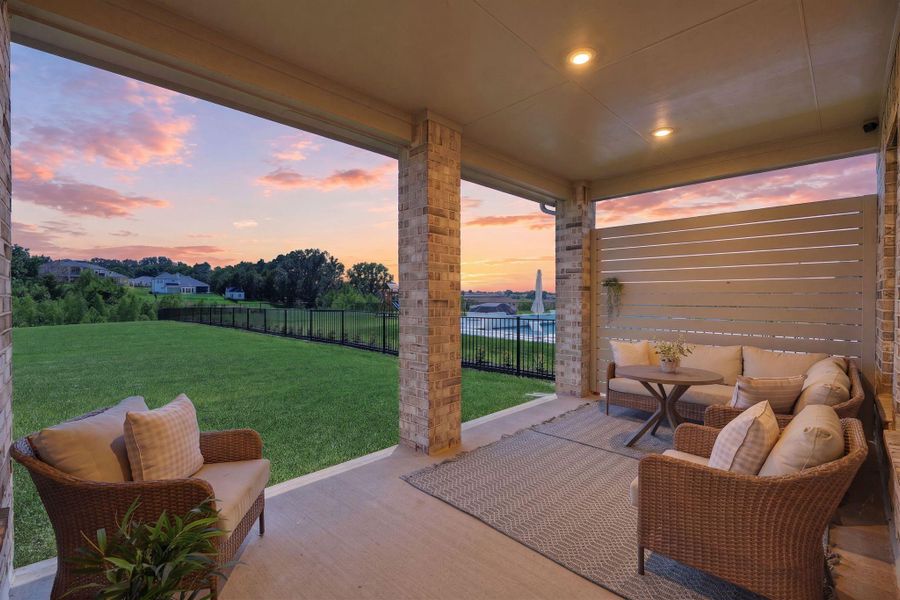This photo showcases a cozy covered patio with wicker seating and a table, offering a relaxing outdoor living space. The area overlooks a spacious, fenced backyard with a scenic view of a lush green landscape and a beautiful sunset sky.