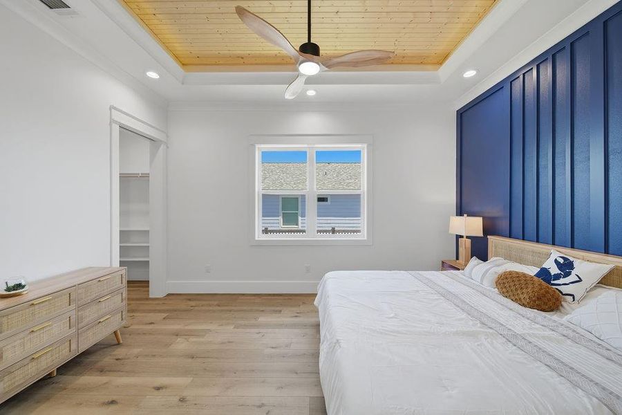 This bedroom features a spacious layout with a light wood floor, a blue accent wall, and a wooden ceiling with a modern fan. It includes a large bed and a dresser, with natural light streaming through the window. There's also an open closet for storage.