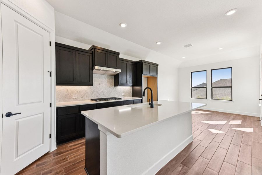 Kitchen featuring backsplash, wood finish floors, light stone counters, recessed lighting, and an island with sink