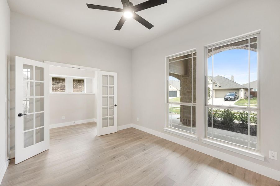 Empty room with light wood floors, ceiling fan, large windows, and French doors leading to adjacent room