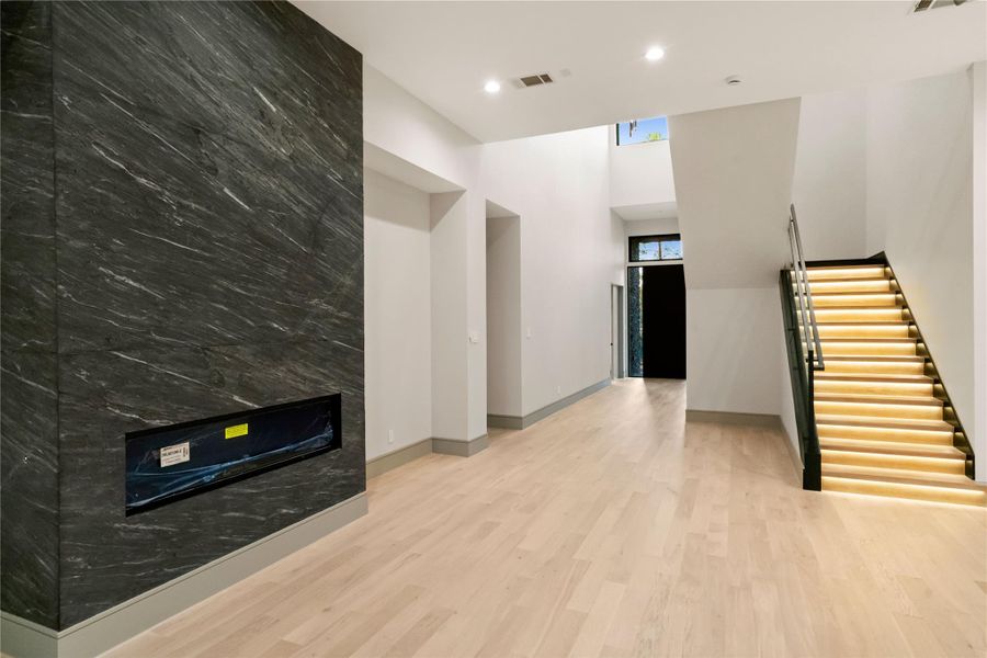 This modern interior features a spacious, high-ceilinged room with light wood flooring and a sleek, dark stone accent wall with a built-in fireplace. The staircase is illuminated with contemporary lighting, enhancing the open, airy feel of the space.