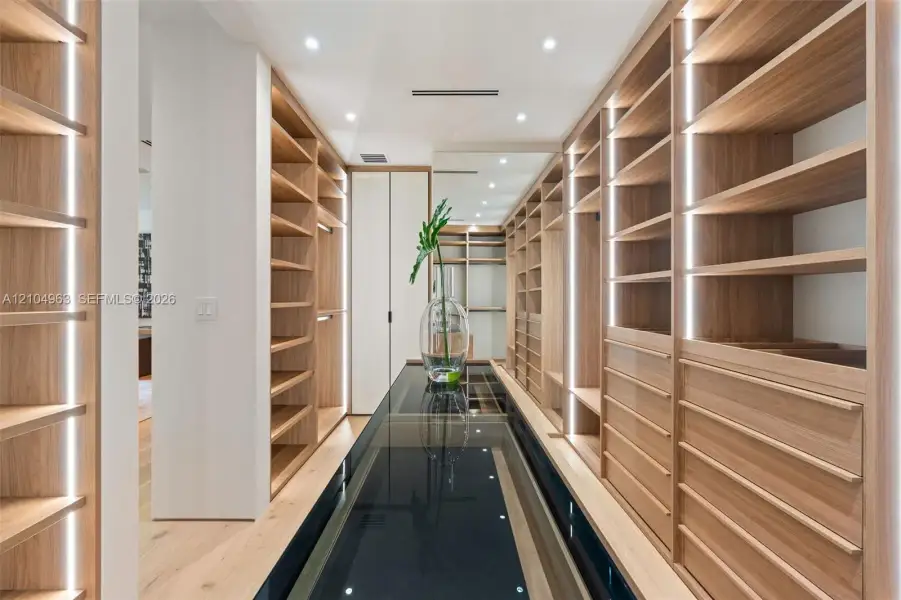 Custom storage cabinetry featuring light wood-finish shelving, integrated vertical LED lighting, a glass-topped center island, and recessed ceiling lighting