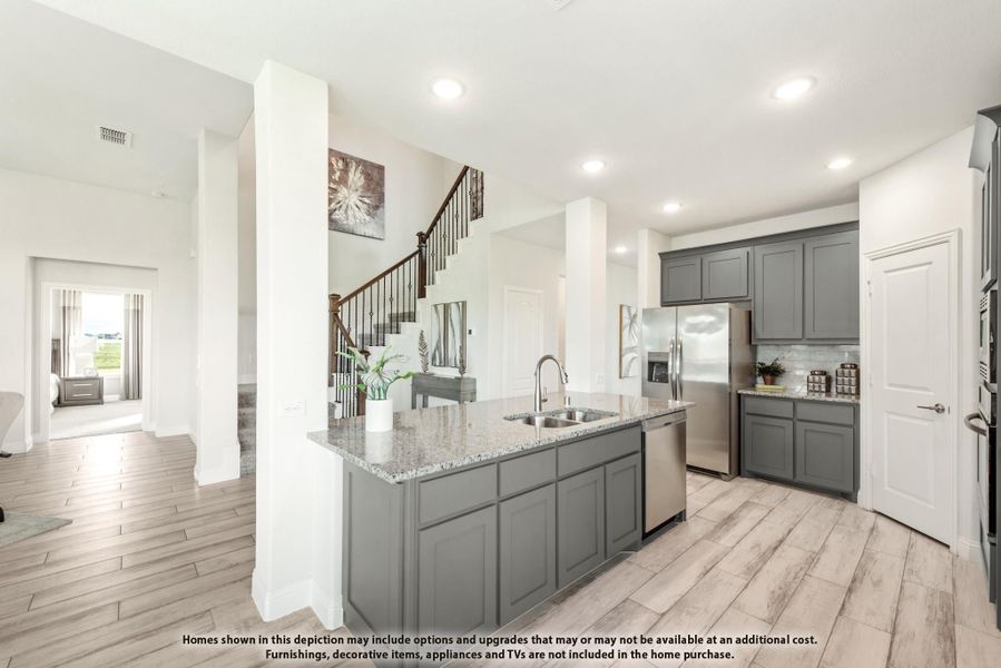 Kitchen with gray cabinets, granite island with sink, stainless appliances, and light wood floors open to staircase entry