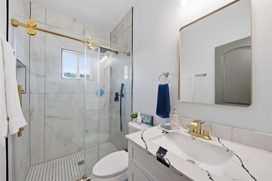This bathroom features a modern design with a sleek marble-look shower, gold fixtures, and a spacious vanity. The glass shower doors and large mirror enhance the bright, airy feel.