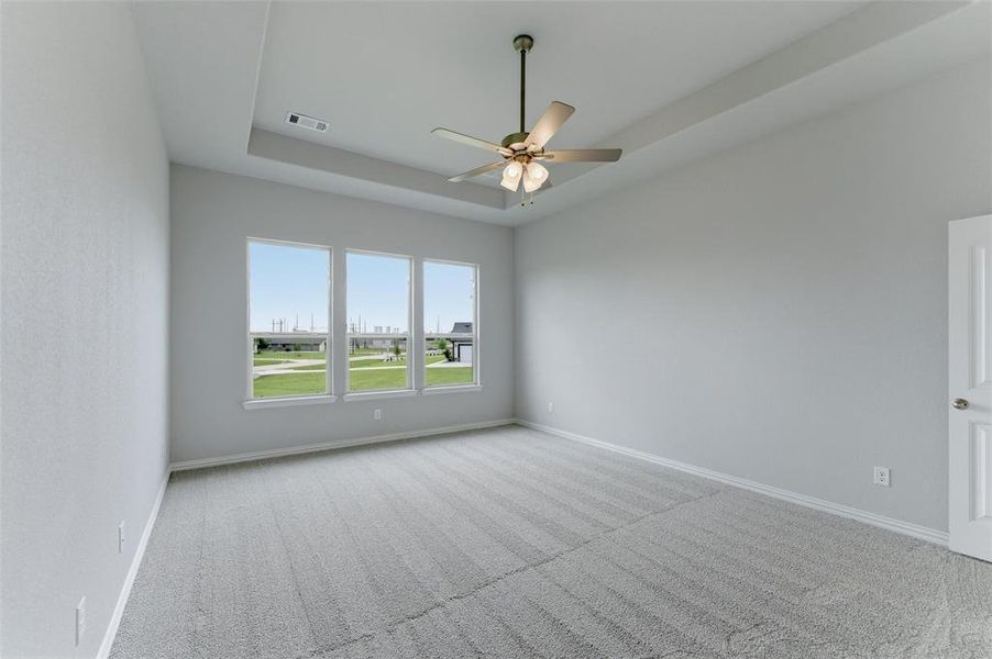 This spacious room features a tray ceiling with a ceiling fan, three windows providing natural light, and carpeting