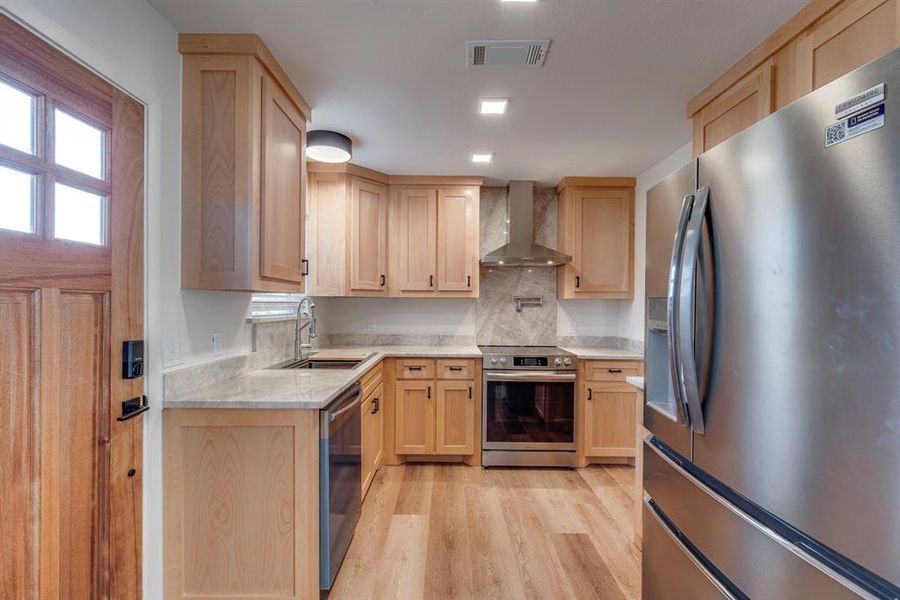 Kitchen with wall chimney exhaust hood, sink, light brown cabinets, stainless steel appliances, and light hardwood / wood-style floors