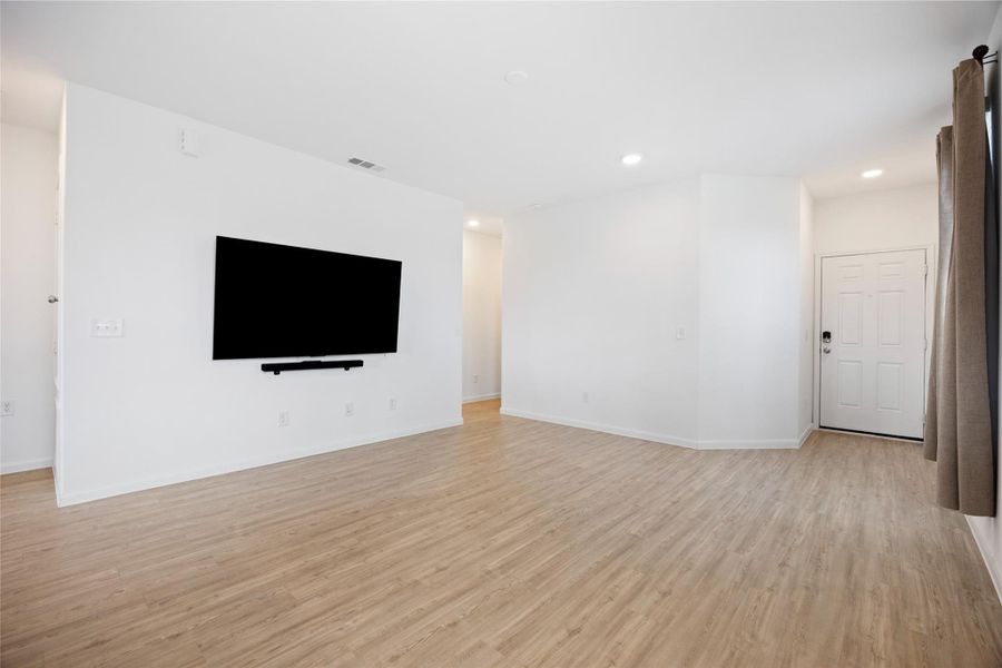 Bright, open living space with light wood flooring, white walls, and a mounted TV. Recessed lighting adds modern touches, and a window with curtains brings in natural light. The entryway features a secure door with a keypad lock.