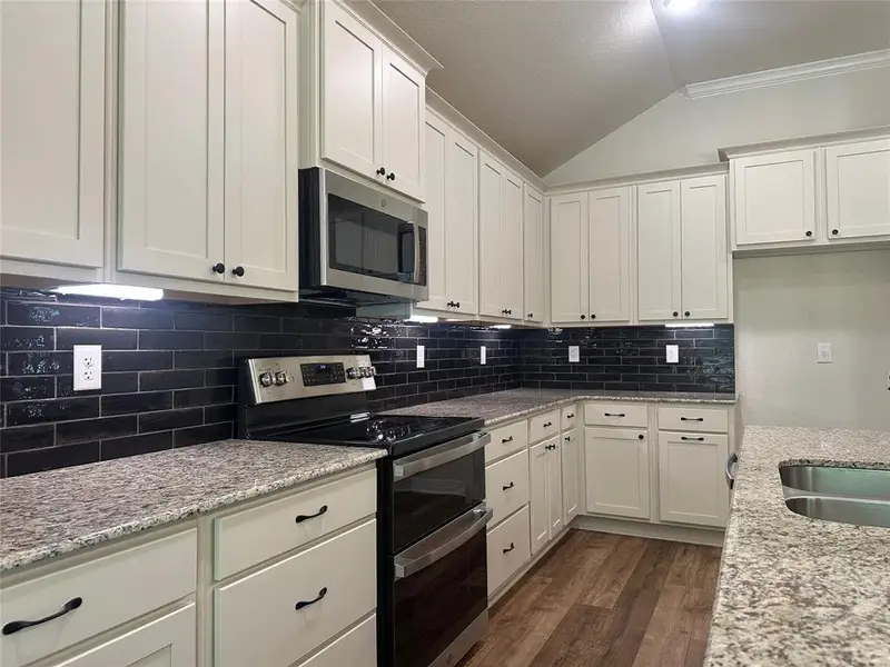 Kitchen with appliances with stainless steel finishes, vaulted ceiling, dark wood-style flooring, white cabinetry, and light stone countertops Kitchen with appliances with stainless steel finishes, vaulted ceiling, dark wood-style flooring, white cabinetry, and light stone countertops