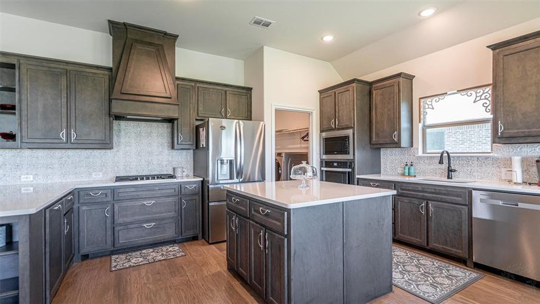 Kitchen featuring a large central island with a light countertop, wood-finish flooring, full-height cabinetry, stainless steel appliances, and a mosaic tile backsplash