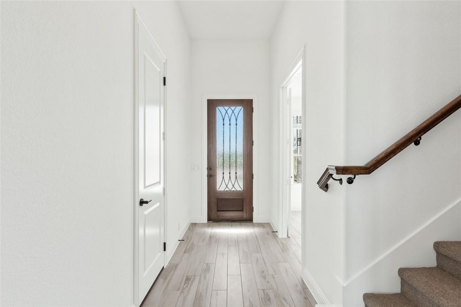 Bright entryway hallway with wood front door, decorative glass panel, light hardwood floors, and staircase with wood railing. Bright entryway hallway with wood front door, decorative glass panel, light hardwood floors, and staircase with wood railing.