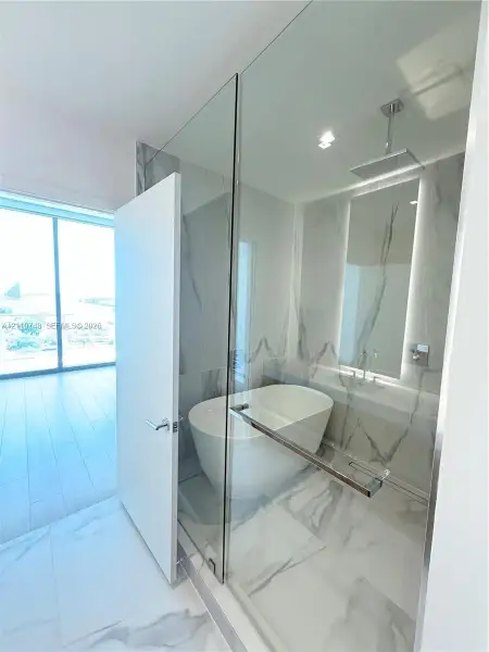 Freestanding soaking tub enclosed in a glass-partitioned wet room featuring floor-to-ceiling stone-look tiling, a ceiling-mounted rainfall showerhead, and a wall-mounted faucet