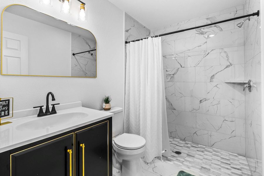 This bathroom features elegant tile, a sleek black vanity with gold accents, a large mirror, and modern fixtures. The shower area is spacious with a curtain and contemporary design.