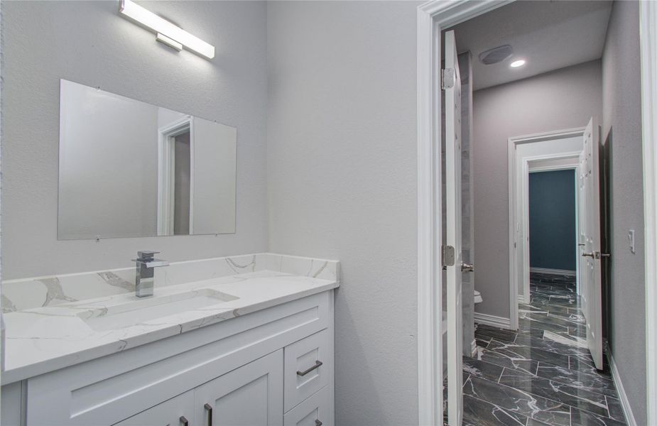 This photo showcases a modern bathroom with a sleek white vanity, marble countertop, and a large mirror. The space features light gray walls and dark, marble-patterned flooring that extends into a hallway with multiple doors, adding to the home's contemporary design.