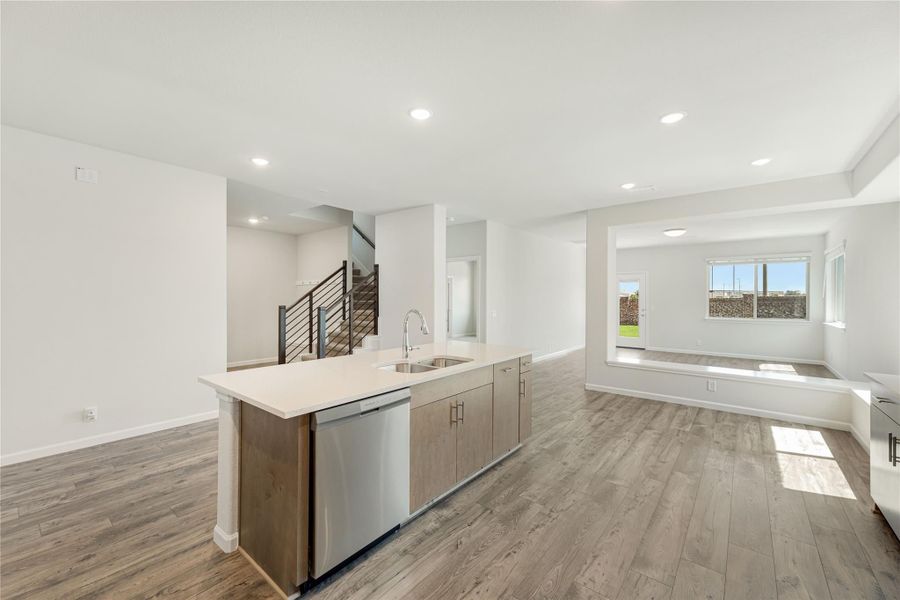 Open-concept kitchen with island sink, stainless dishwasher, wood-look floors, and living area with staircase visible.