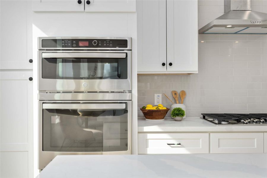 A sleek stainless steel double-oven is built into bright white cabinetry and set beside a stylish glossy subway-tile backsplash, with polished white quartz countertops and simple decor adding to the kitchen’s clean, modern look.