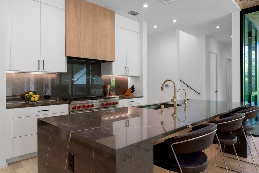 This custom crafted gourmet kitchen boasts a large center island wrapped in high-end waterfall marble countertops, which extend throughout the kitchen and wrap up behind the gas range wall.