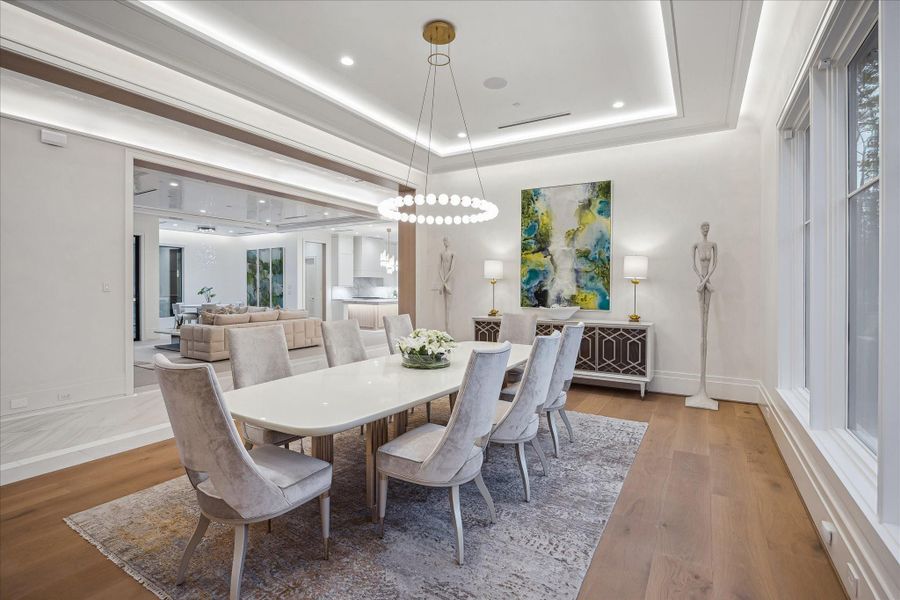 Elegant dining room featuring Venetian plaster walls and a layered tray ceiling with LED cove lighting and recessed fixtures. Wide-plank white oak wood flooring adds warmth, while large picture windows fill the space with natural light.