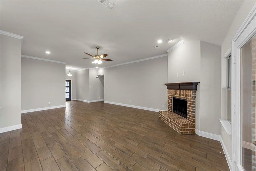 Expansive open-concept living area featuring wood-finish flooring, a brick fireplace with a dark wood mantel, recessed lighting, and crown molding