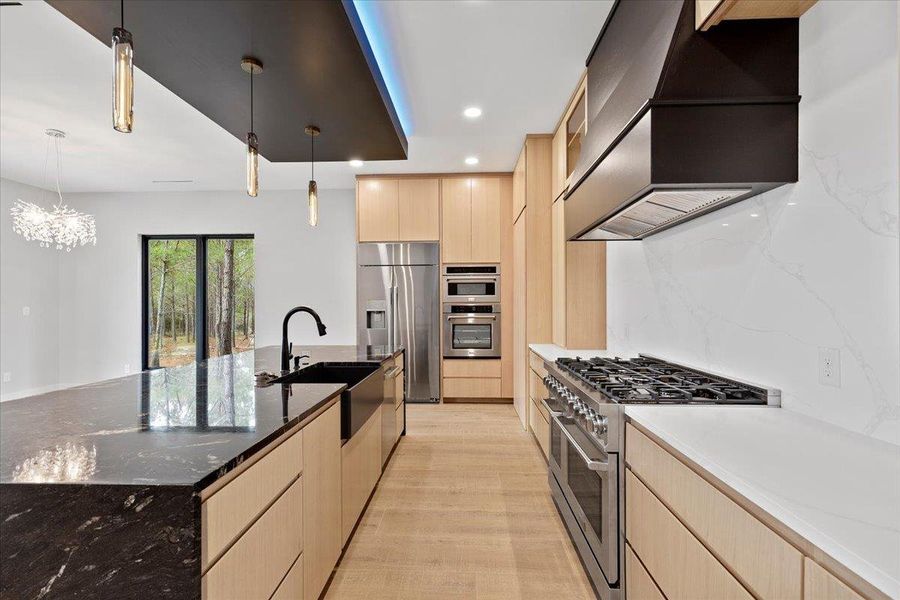 Kitchen featuring light wood cabinets, dark natural stone granite countertops, premium appliances, custom range hood, and a large island with basin sink. Kitchen featuring light wood cabinets, dark natural stone granite countertops, premium appliances, custom range hood, and a large island with basin sink.
