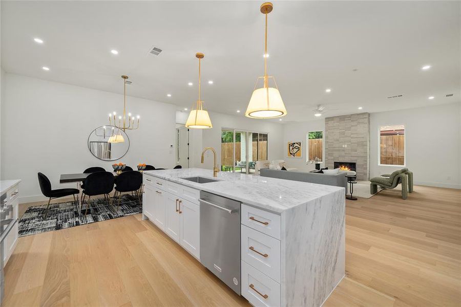 This open-concept living space features hardwood flooring throughout, a kitchen island with a marble countertop, white cabinetry with bronze hardware, and a stainless steel dishwasher