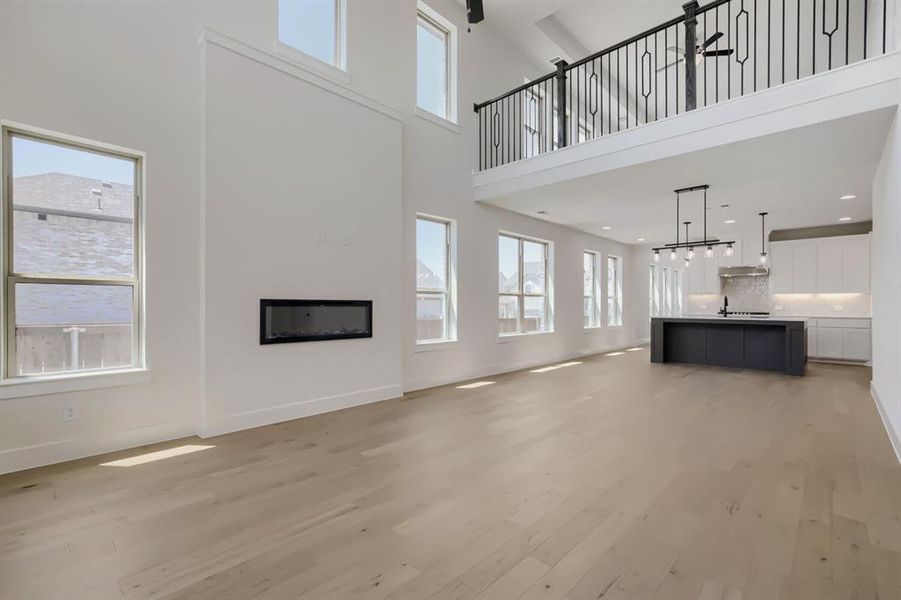 Open-concept living area featuring light wood-finish flooring, a wall-mounted linear fireplace, and an expansive kitchen with a dark-toned island and light cabinetry