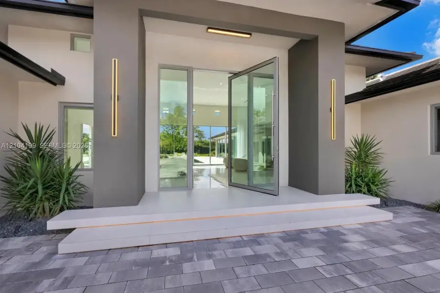 Modern entryway featuring an oversized glass pivot door, dual tiered concrete steps, linear wall-mounted exterior lighting, and a paved stone driveway