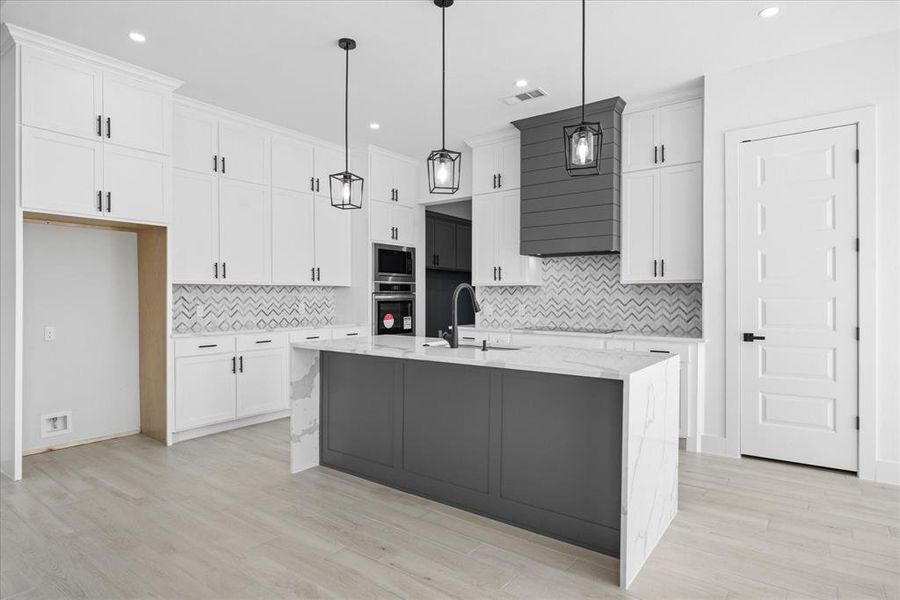 Kitchen featuring a waterfall island with stone countertop, white shaker cabinetry, patterned tile backsplash, built-in double oven, and wood-finish flooring