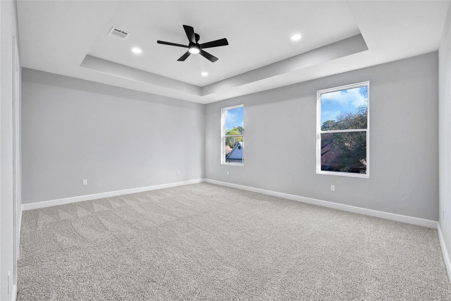 Spacious bedroom with soft carpet flooring, two large windows for natural light, and a modern ceiling fan. Features a stylish tray ceiling and recessed lighting, creating a bright and inviting atmosphere.