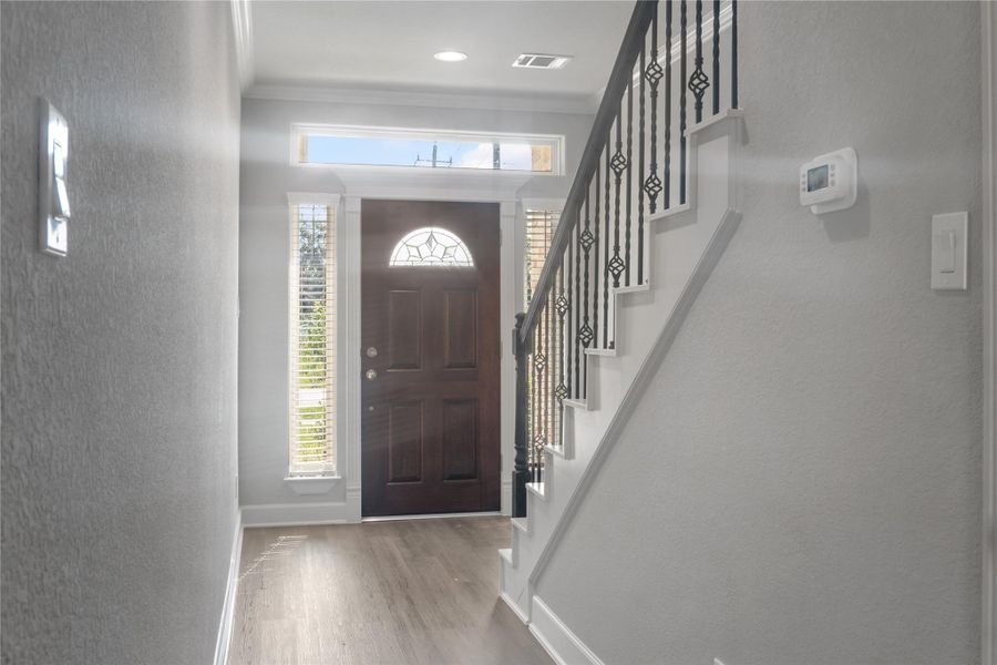 Bright entryway with a dark wood front door and sidelights, leading to a staircase with stylish black iron railings. Light gray walls and wood flooring create a welcoming atmosphere.