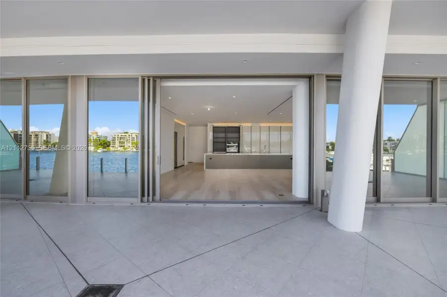 Covered terrace featuring stone tile flooring and a structural cylindrical column, spanning wide sliding glass doors that reveal wood-finish flooring and built-in kitchen cabinetry