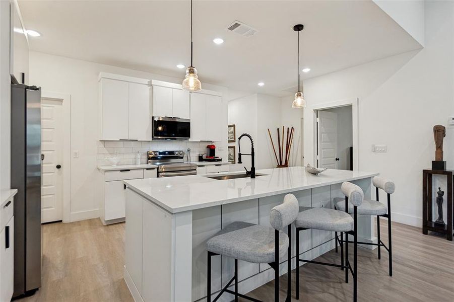 Modern kitchen featuring a large central island with seating, white shaker cabinetry, stainless steel appliances, and wood-finish flooring Modern kitchen featuring a large central island with seating, white shaker cabinetry, stainless steel appliances, and wood-finish flooring