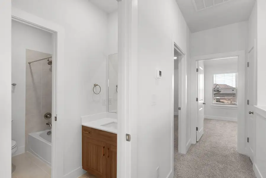 A pristine bathroom features a crisp white vanity and a shower-tub combination, with soft carpeting leading to an adjacent bedroom.