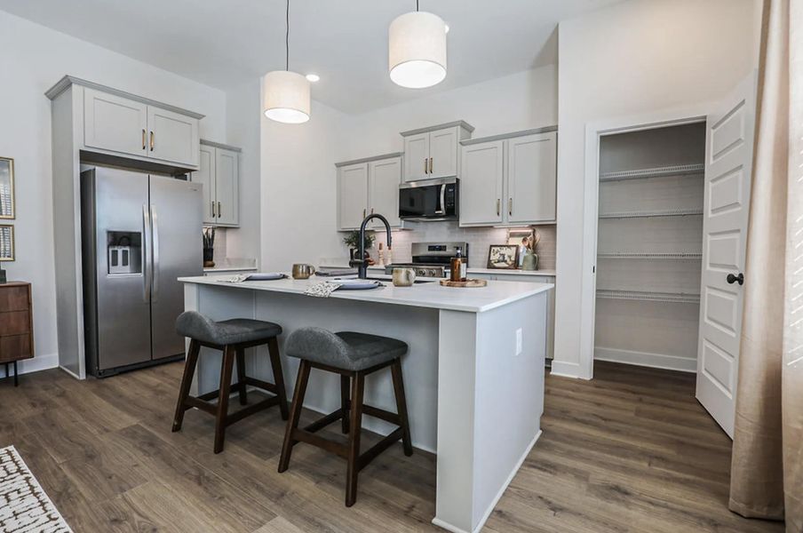 Modern kitchen featuring a central island with countertop seating, light grey cabinetry, stainless steel appliances, wood-finish flooring, and a spacious pantry with wire shelving