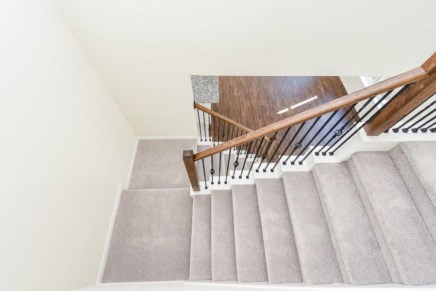 A top-down view of a carpeted staircase with wooden handrails and black metal balusters, leading to a hardwood floor landing.