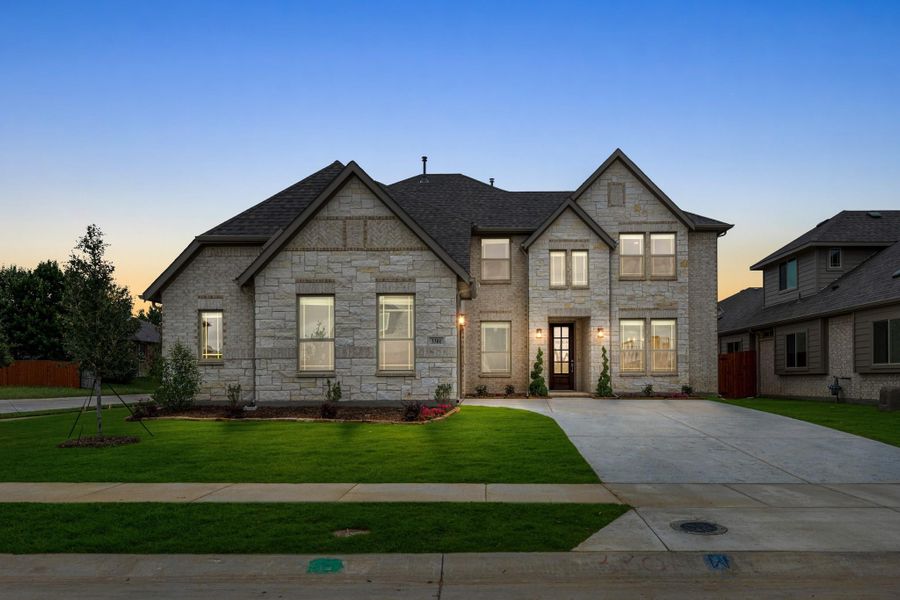 Two-story home with light stone exterior, dark roof, front entry door, and manicured lawn at dusk