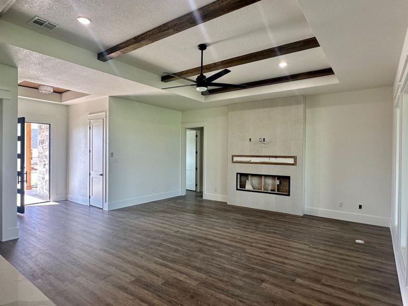 Unfurnished living room with ceiling fan, a tiled fireplace, dark wood finished floors, beam ceiling, and a tray ceiling