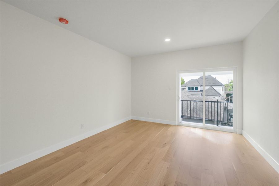 Bright and spacious room with light wood flooring and neutral walls, featuring a sliding glass door that opens onto a balcony with a view of neighboring rooftops. Perfect for a bedroom or home office.