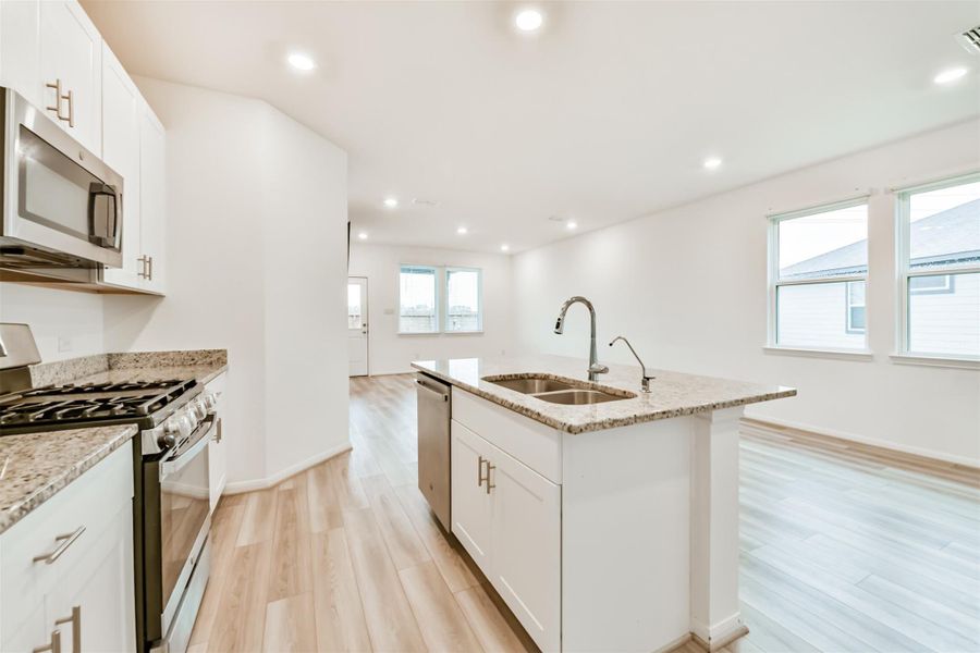 Open-concept kitchen with white cabinetry, granite countertops, and stainless steel appliances. The kitchen island features a sink and ample workspace, and the area is well-lit with recessed lighting and natural light from large windows.