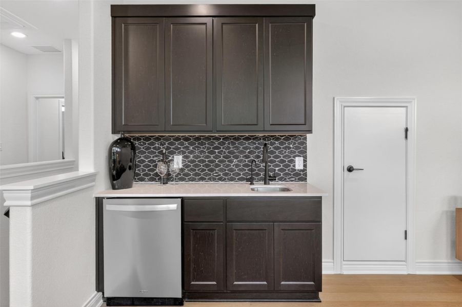 A refined dry bar with dark cabinetry, stylish hexagonal tile backsplash, and a sleek stainless steel dishwasher offers seamless entertaining. A refined dry bar with dark cabinetry, stylish hexagonal tile backsplash, and a sleek stainless steel dishwasher offers seamless entertaining.